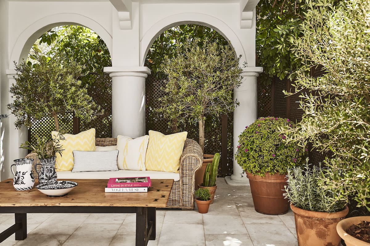A covered outdoor seating area with a sofa, cushions, a table with decorative items, and potted plants.