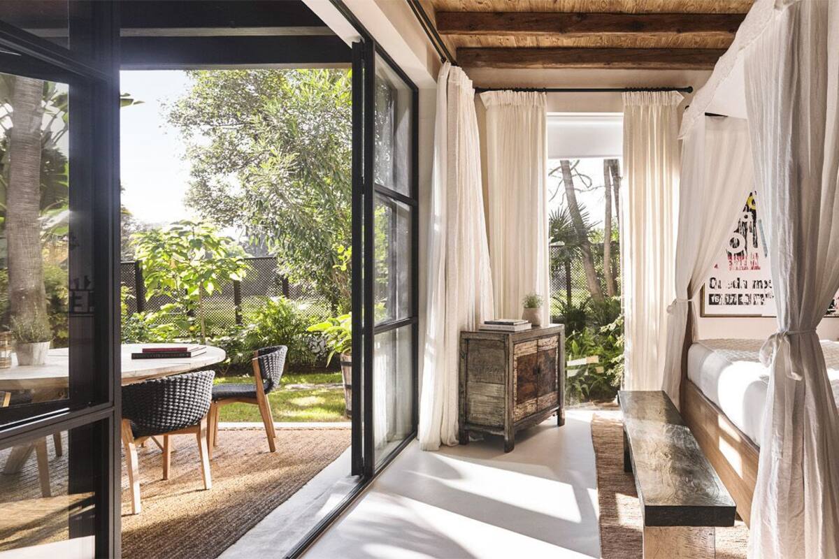 A bedroom with a bed, curtains, a bedside table, and a view of the outdoors.