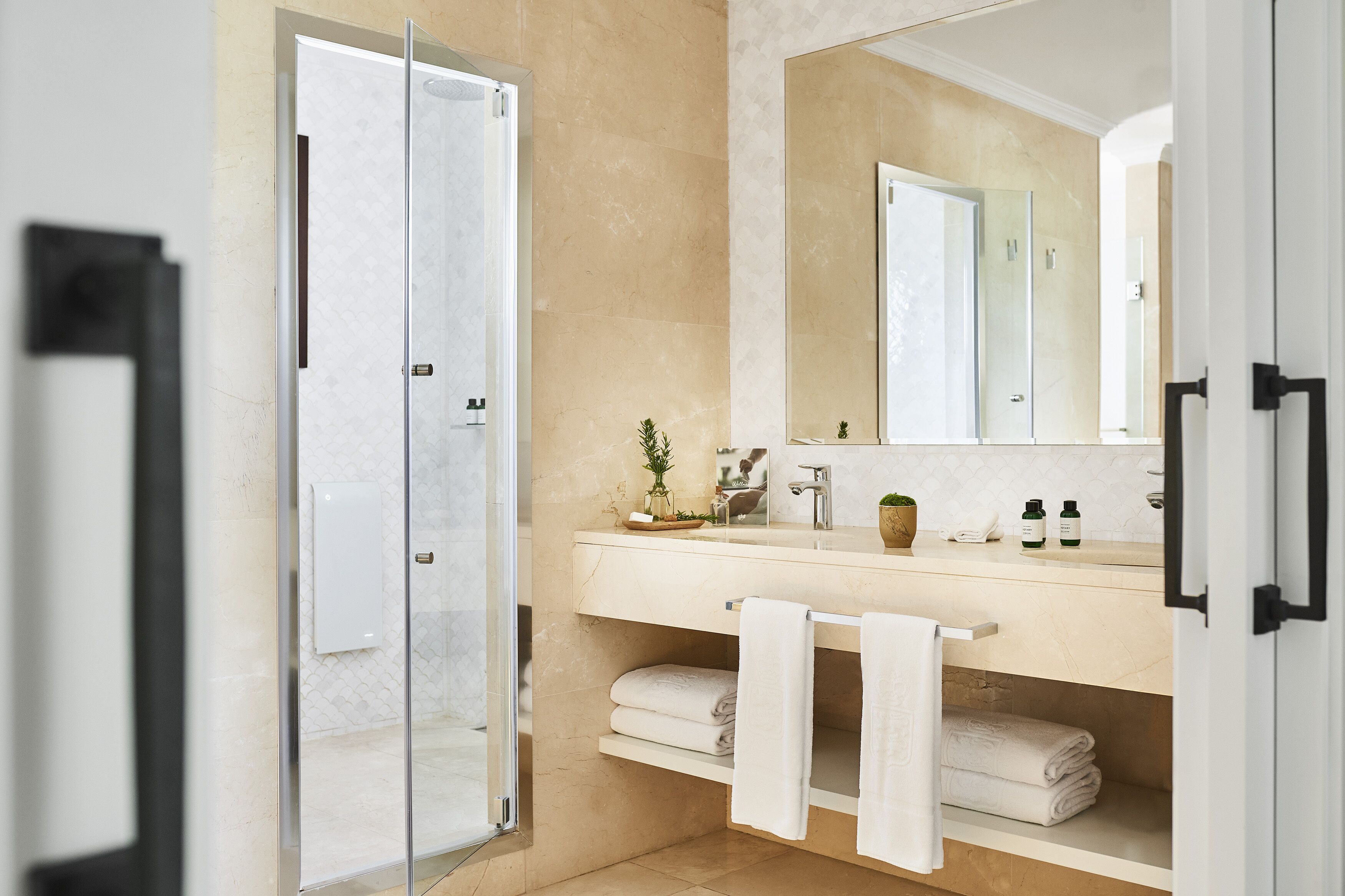 A modern bathroom with a glass shower door, a beige tiled wall, a white sink with a mirror, and neatly folded towels.