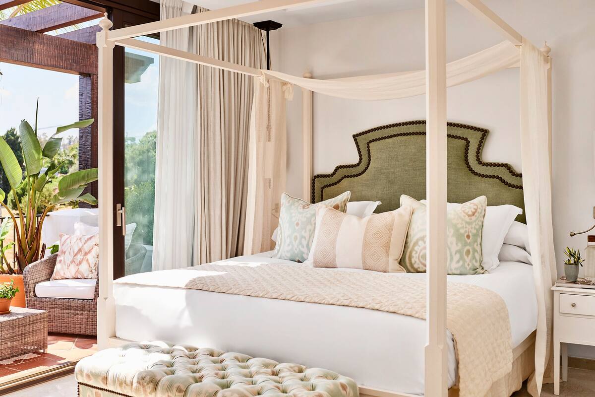 A four-poster canopy bed with a tufted ottoman, a nightstand, and a potted plant.