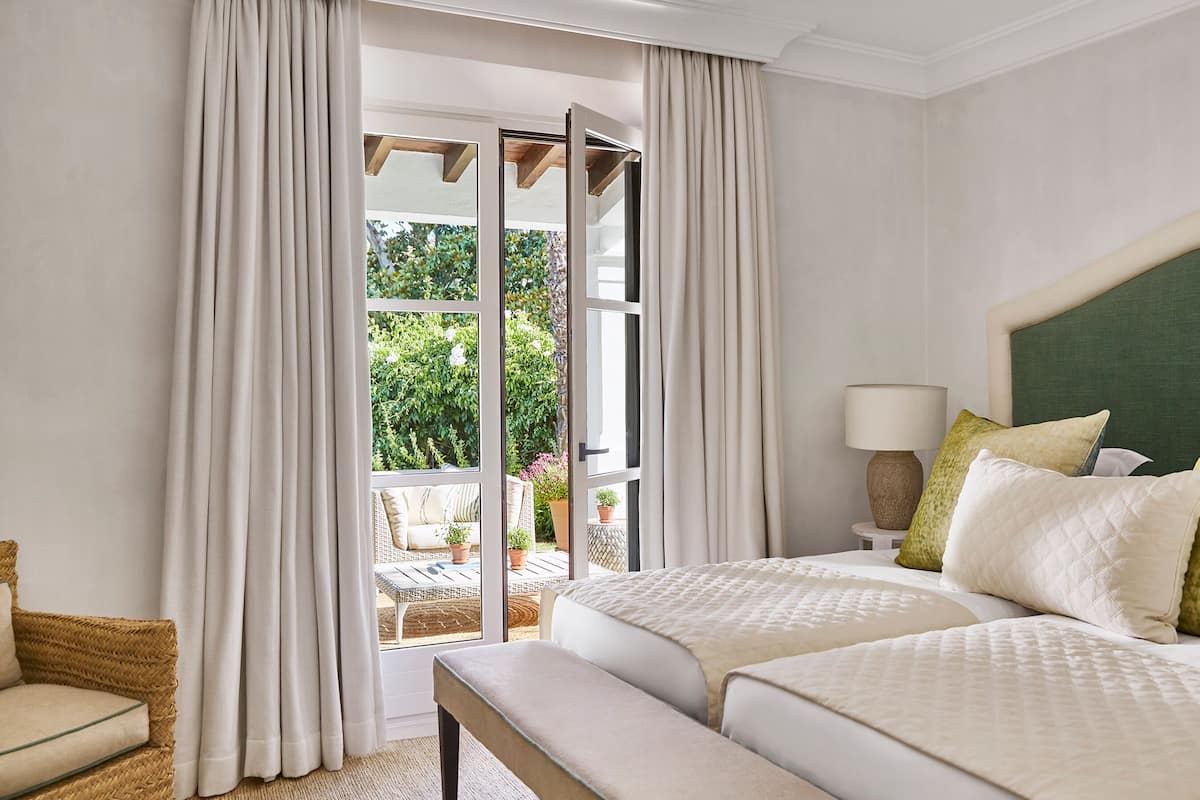 A hotel room with a large bed, a bench, a chair, and a view of a garden through a glass door.
