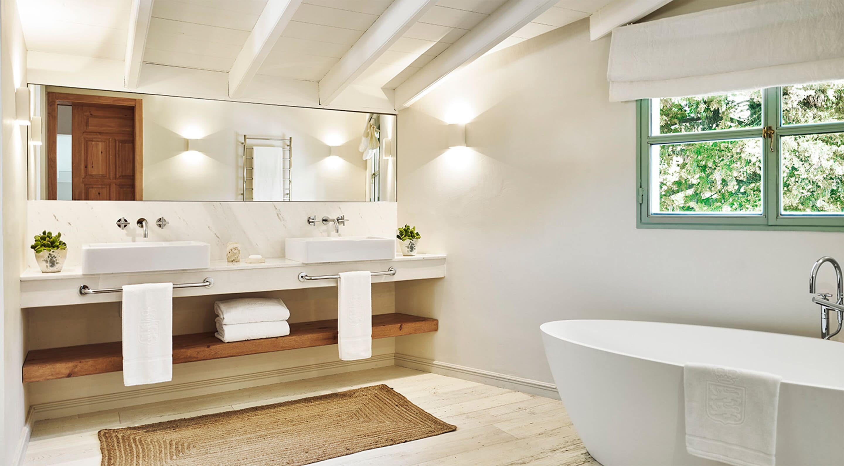 A modern bathroom with two sinks, a large bathtub, a mirror, and a window with a view of greenery.