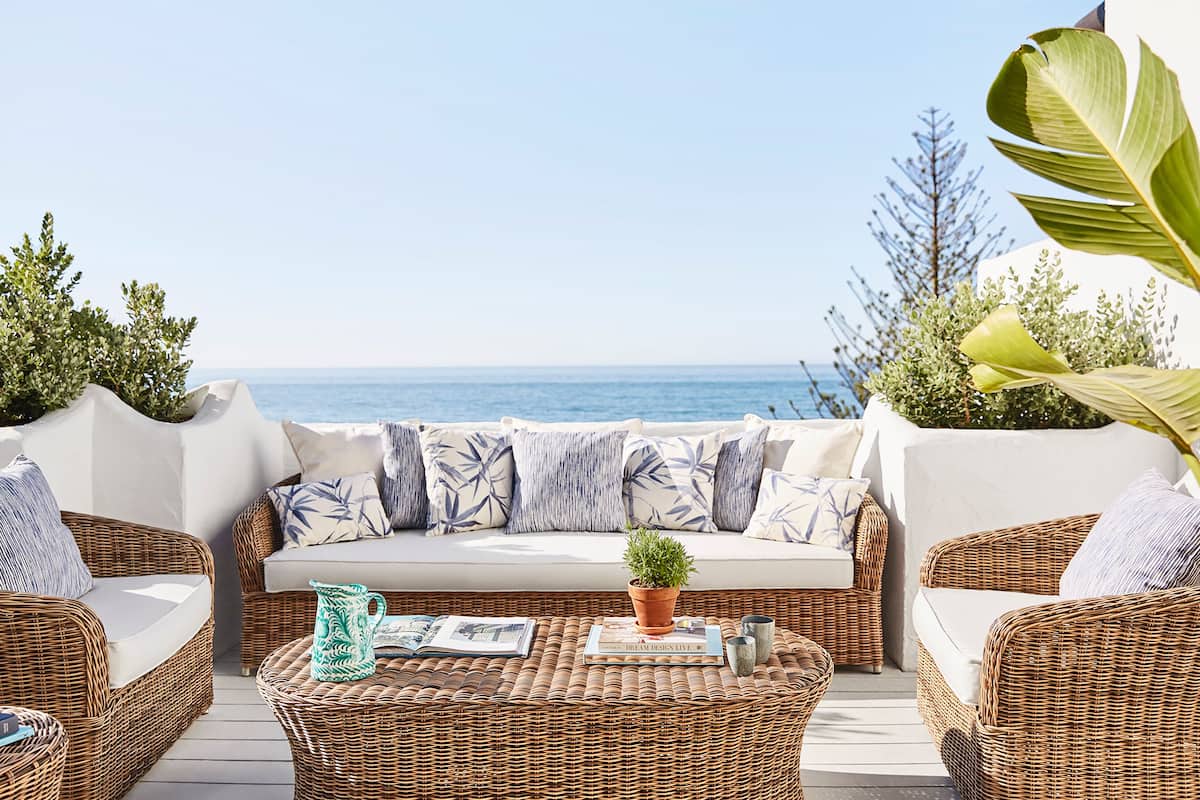 A wicker sofa with cushions, a coffee table with magazines, and a view of the sea.