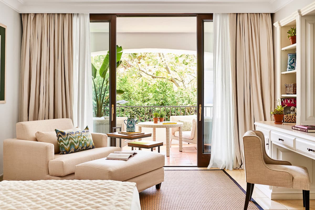 A hotel room with a sofa, armchair, and a desk with a chair. There is a balcony with a view of greenery.