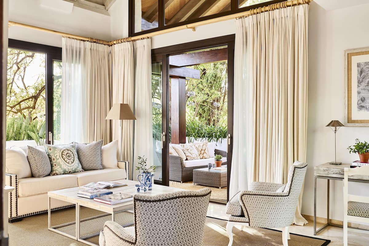 A well-lit living room with a sofa, armchairs, a coffee table, and a dining table. There is a view of greenery through large windows and a door leading outside.