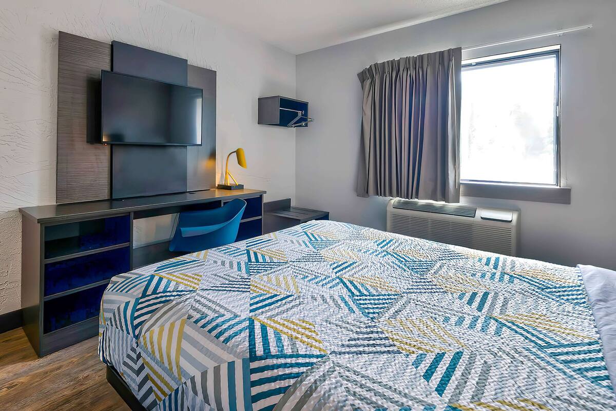 A modern hotel room with a large bed, a desk with a chair, a flat-screen TV mounted on the wall, and a window with curtains.