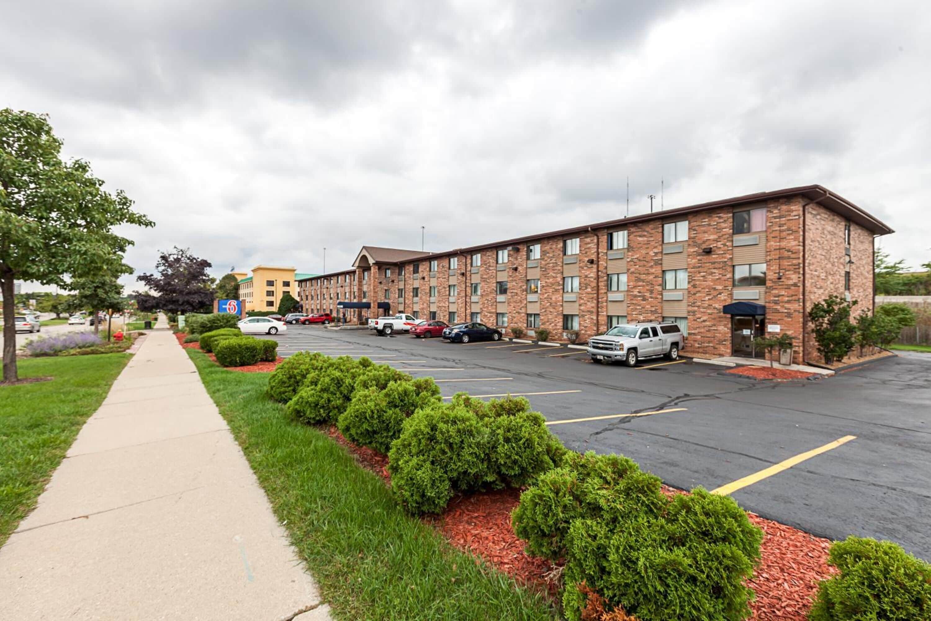 A multi-story hotel with a brick facade, a parking lot, and a sidewalk lined with greenery.