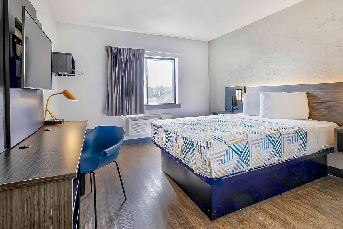 A modern hotel room with a large bed, a desk with a chair, a TV, and a window with curtains.