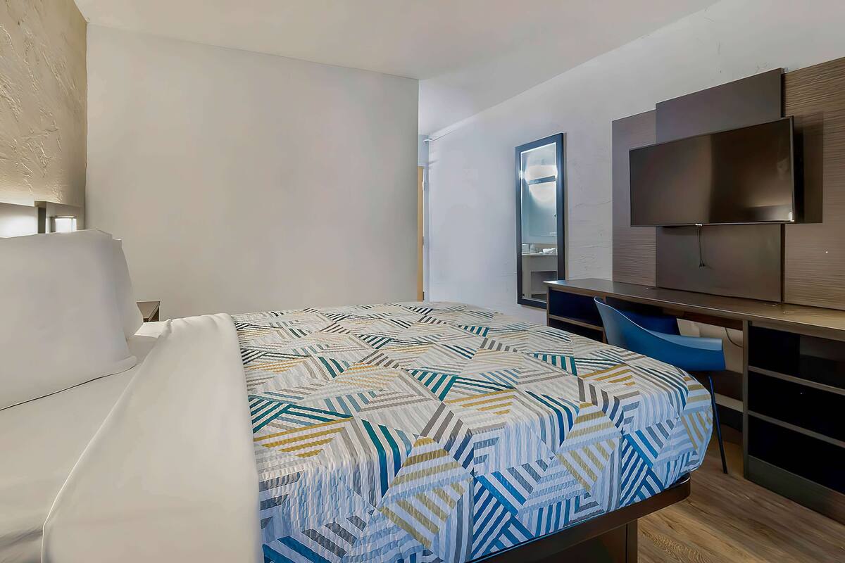 A hotel room with a bed, a desk, a chair, a TV, and a lamp.