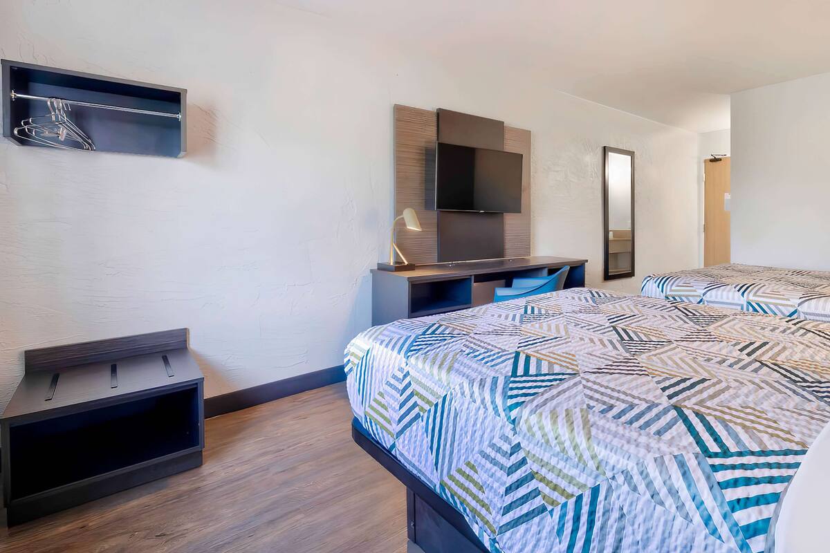 A modern hotel room with a large bed, a desk with a flat-screen TV, a mirror, and a small shelf.