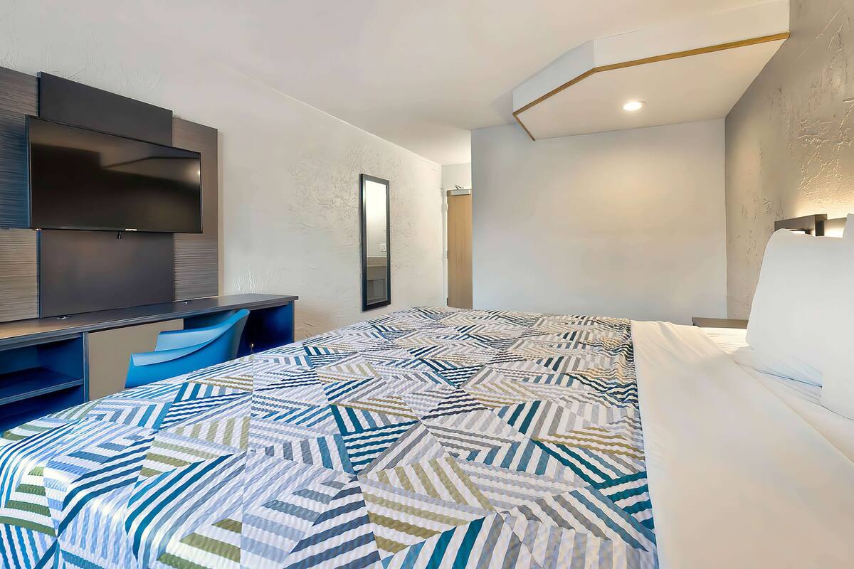 A modern hotel room with a large bed, a desk with a chair, a flat-screen TV, and a large mirror.