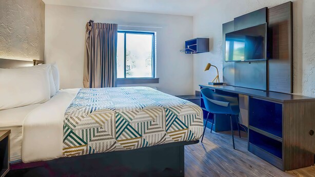 Habitación de hotel con cama, escritorio, silla, televisor y ventana con cortinas.