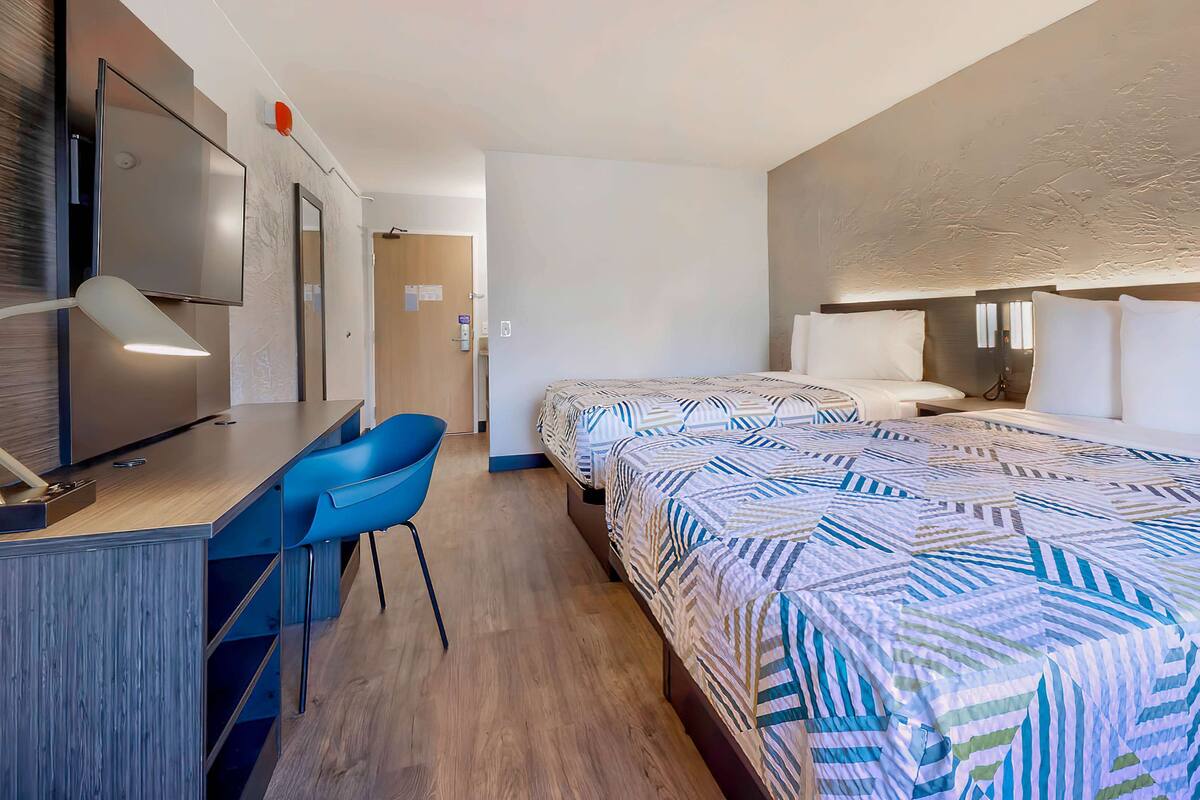 A hotel room with two beds, a desk with a chair, a TV, and a bathroom visible through an open door.