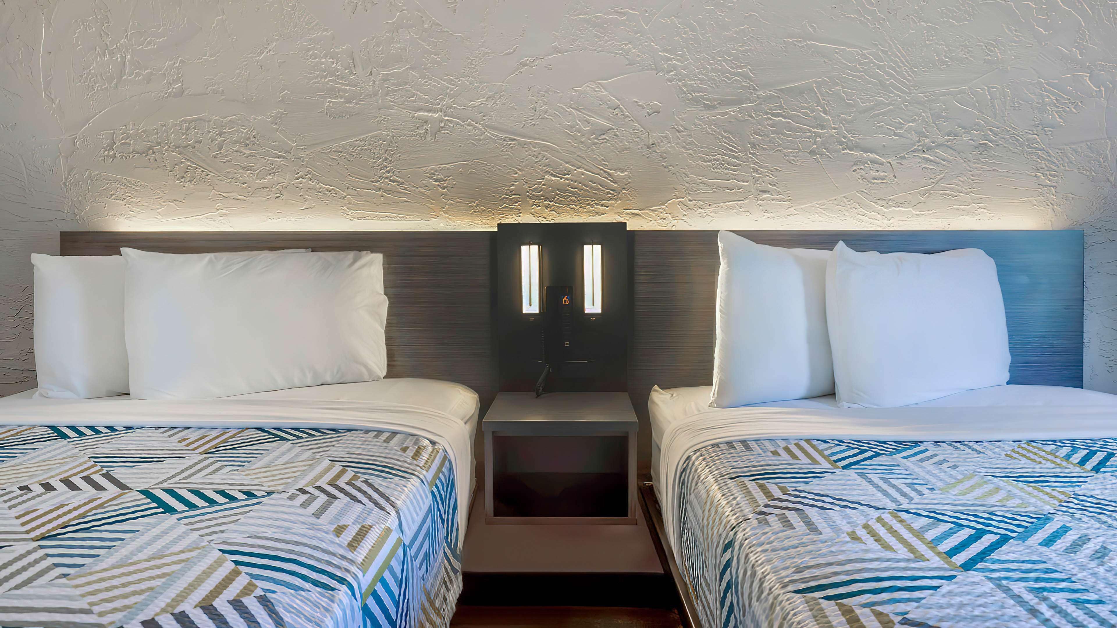 A hotel room with two beds, each with a patterned bedspread, a nightstand, and a wall-mounted light fixture.