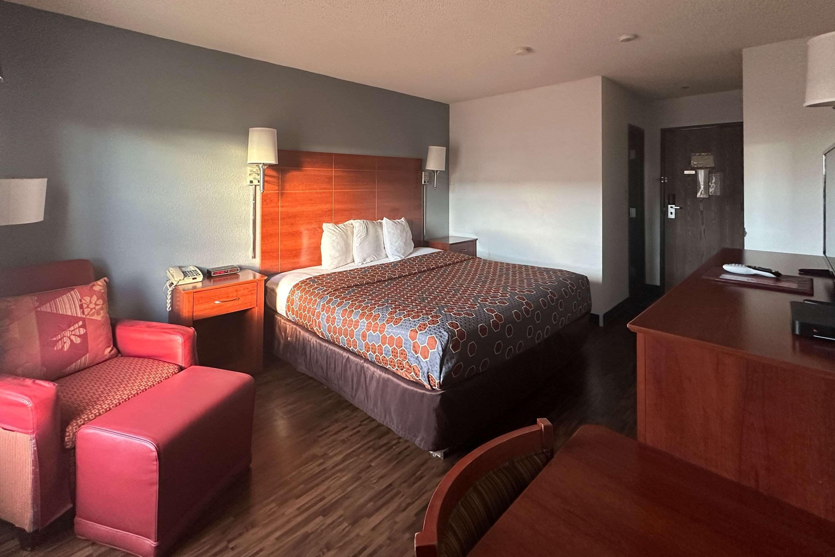 A hotel room with a large bed, a desk, a chair, a bedside table with a lamp, and a wardrobe.