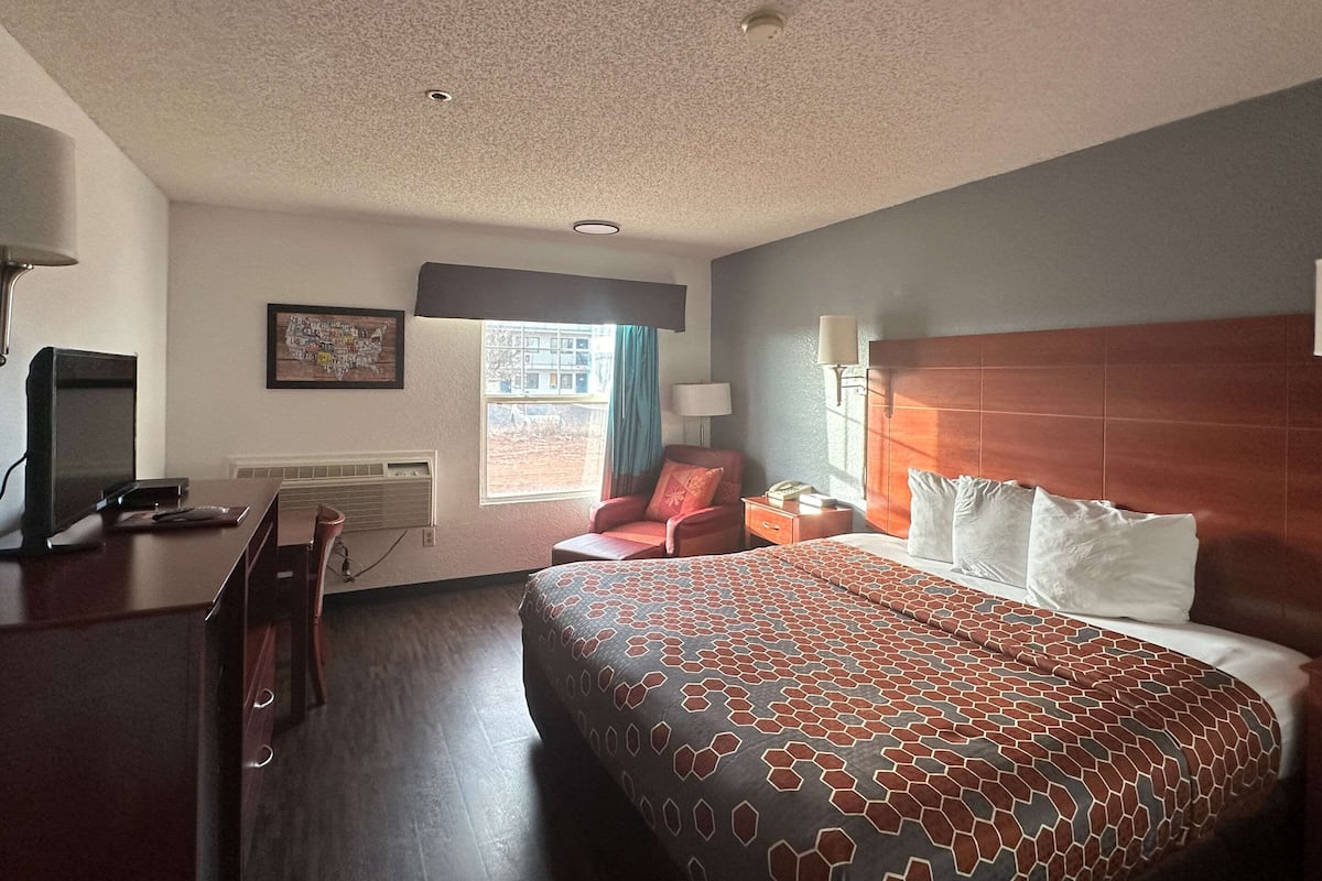 A hotel room with a large bed, a desk with a computer, a chair, a window with curtains, and a wall-mounted lamp.