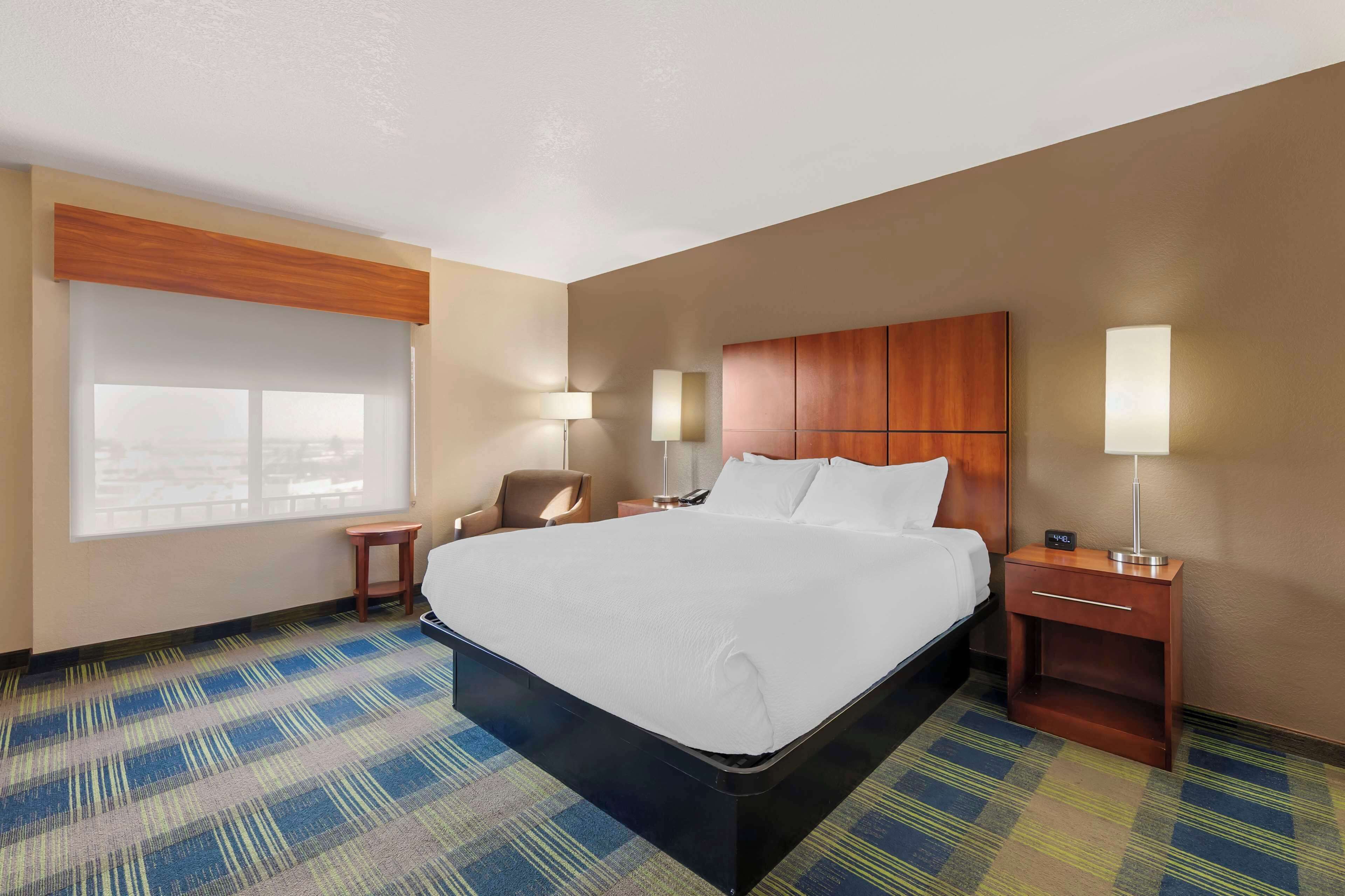 A hotel room with a large bed, bedside tables, a chair, and a window with blinds.