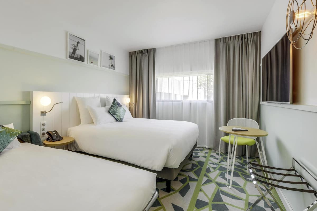 A hotel room with two beds, a small table, a chair, a phone, and a window with curtains.