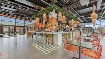 A modern cafe with a central bar, green chairs, and hanging wicker lamps.