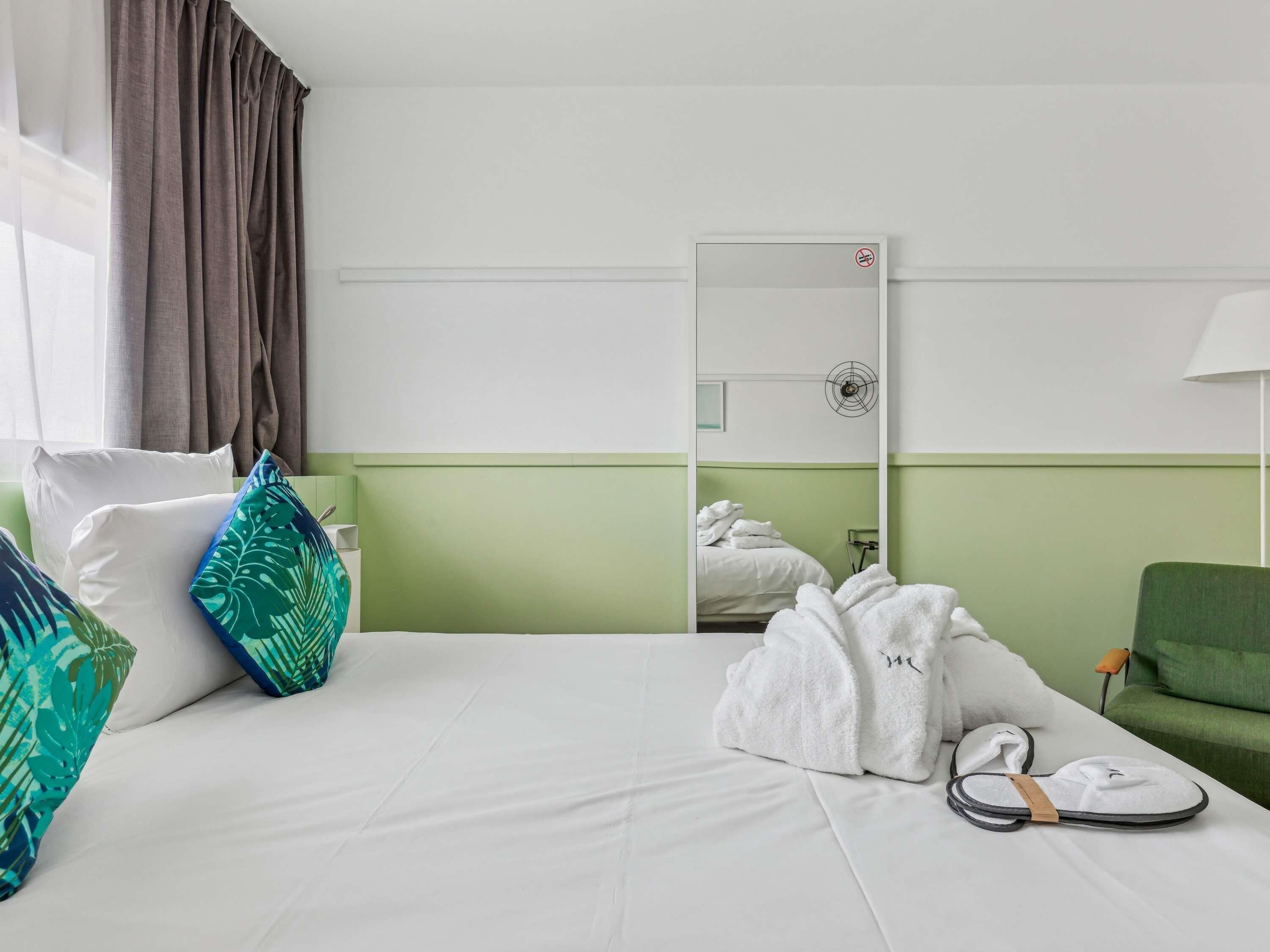 Habitación de hotel con cama, almohadas, un sillón verde, un espejo y una toalla sobre la cama.