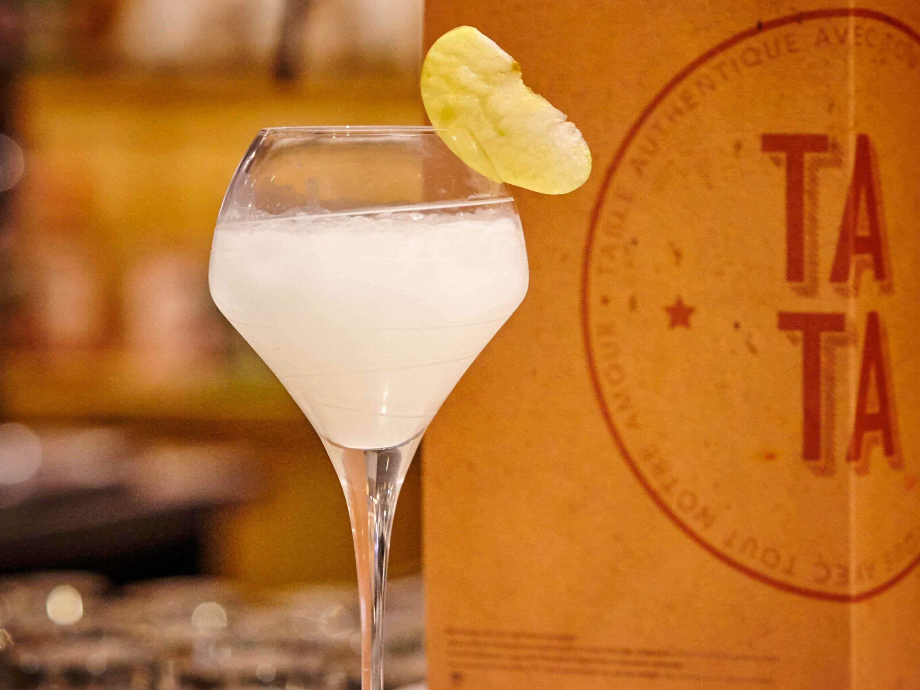 Un verre à martini garni d’une tranche de citron, placé à côté d’un sac en papier brun orné du logo « TATA ».