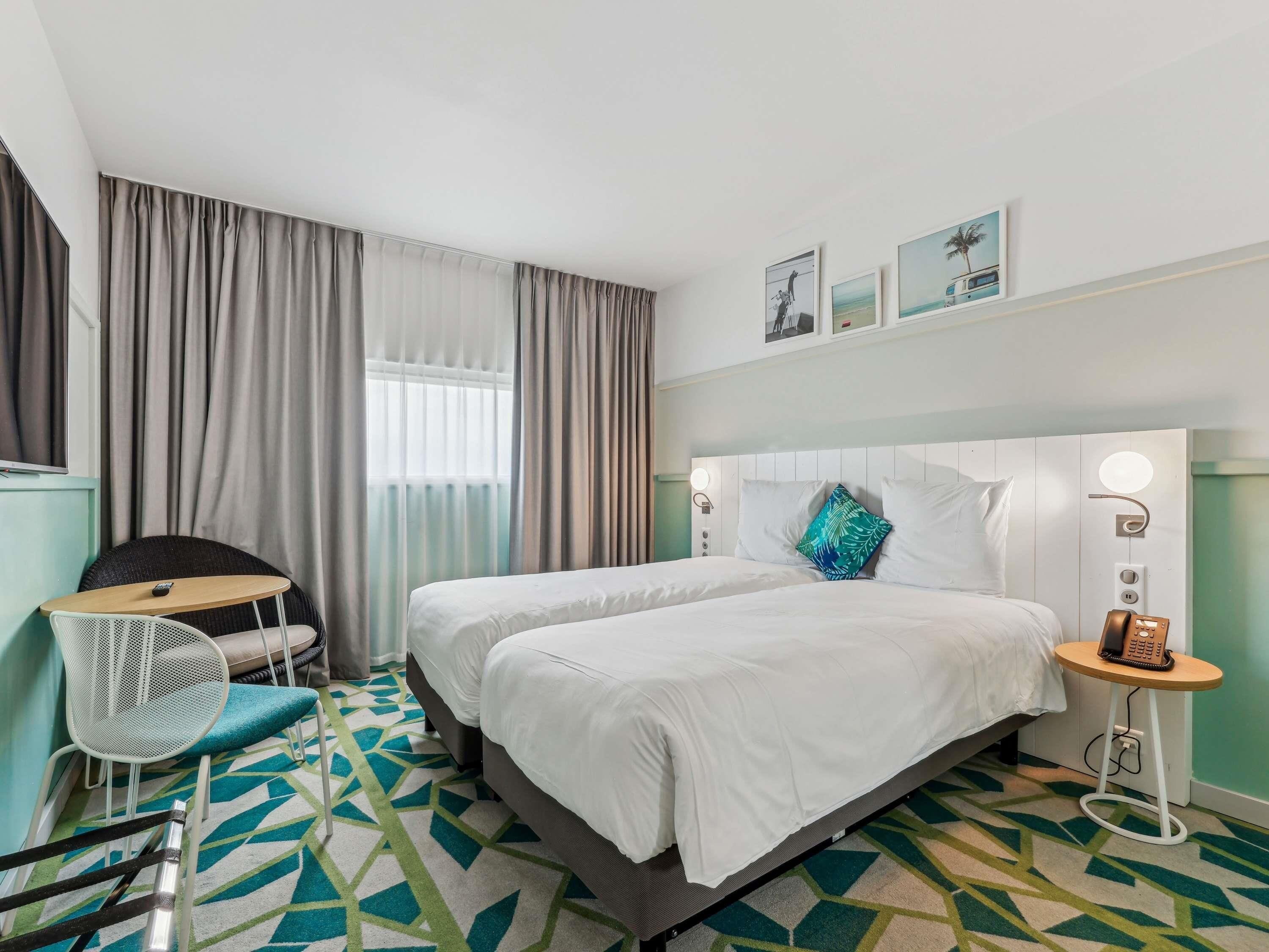 Habitación de hotel con dos camas, un escritorio, una silla, una mesita con un teléfono y una ventana con cortinas.
