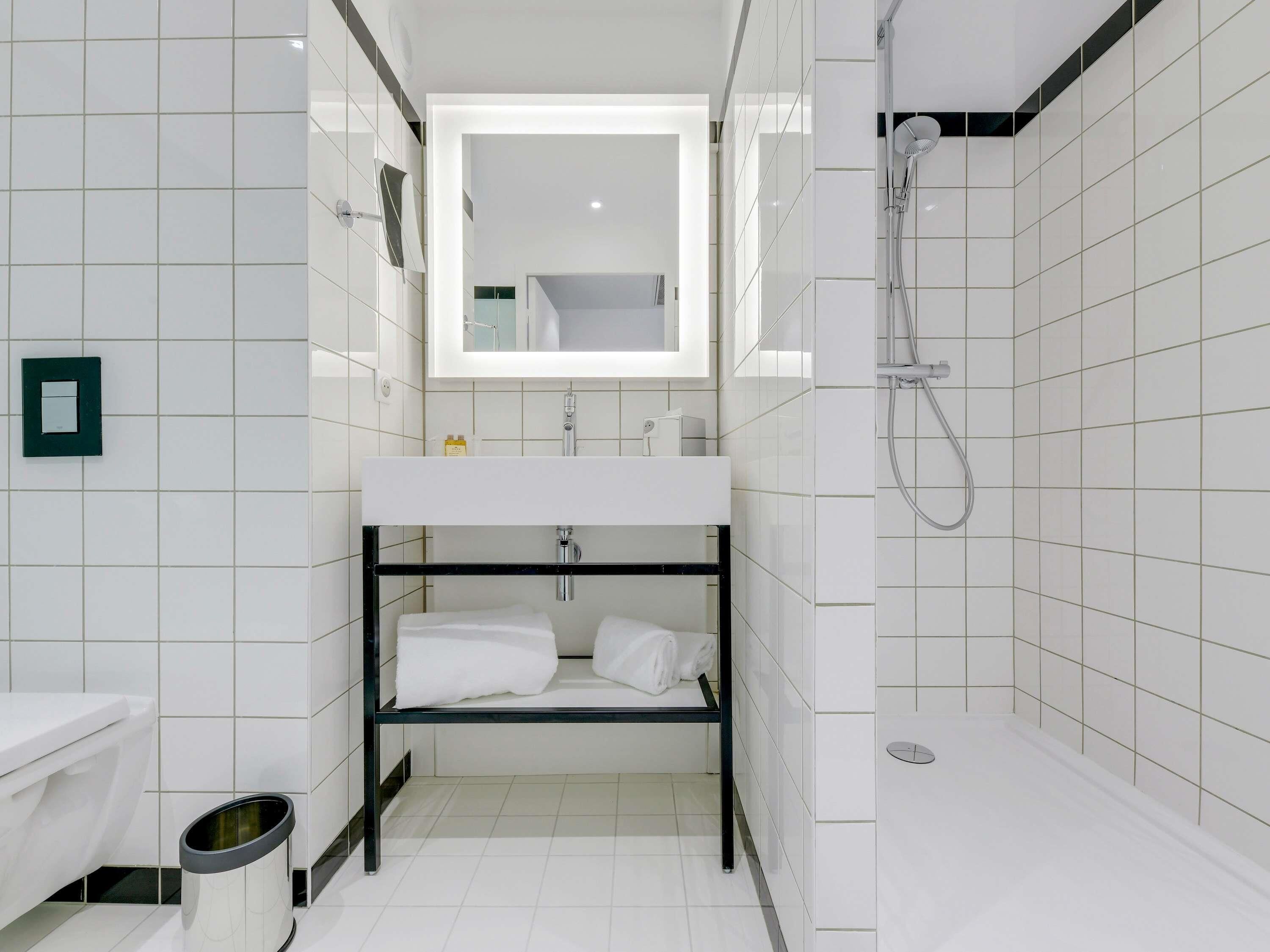 A modern bathroom with a white tiled wall, a white sink with a mirror above it, a black metal shelf with towels, and a shower area.