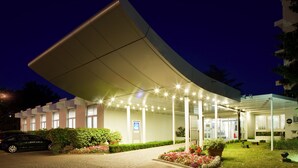A modern hotel entrance with a covered walkway, a car parked outside, and well-maintained landscaping. - Novotel Massy Palaiseau (Palaiseau)