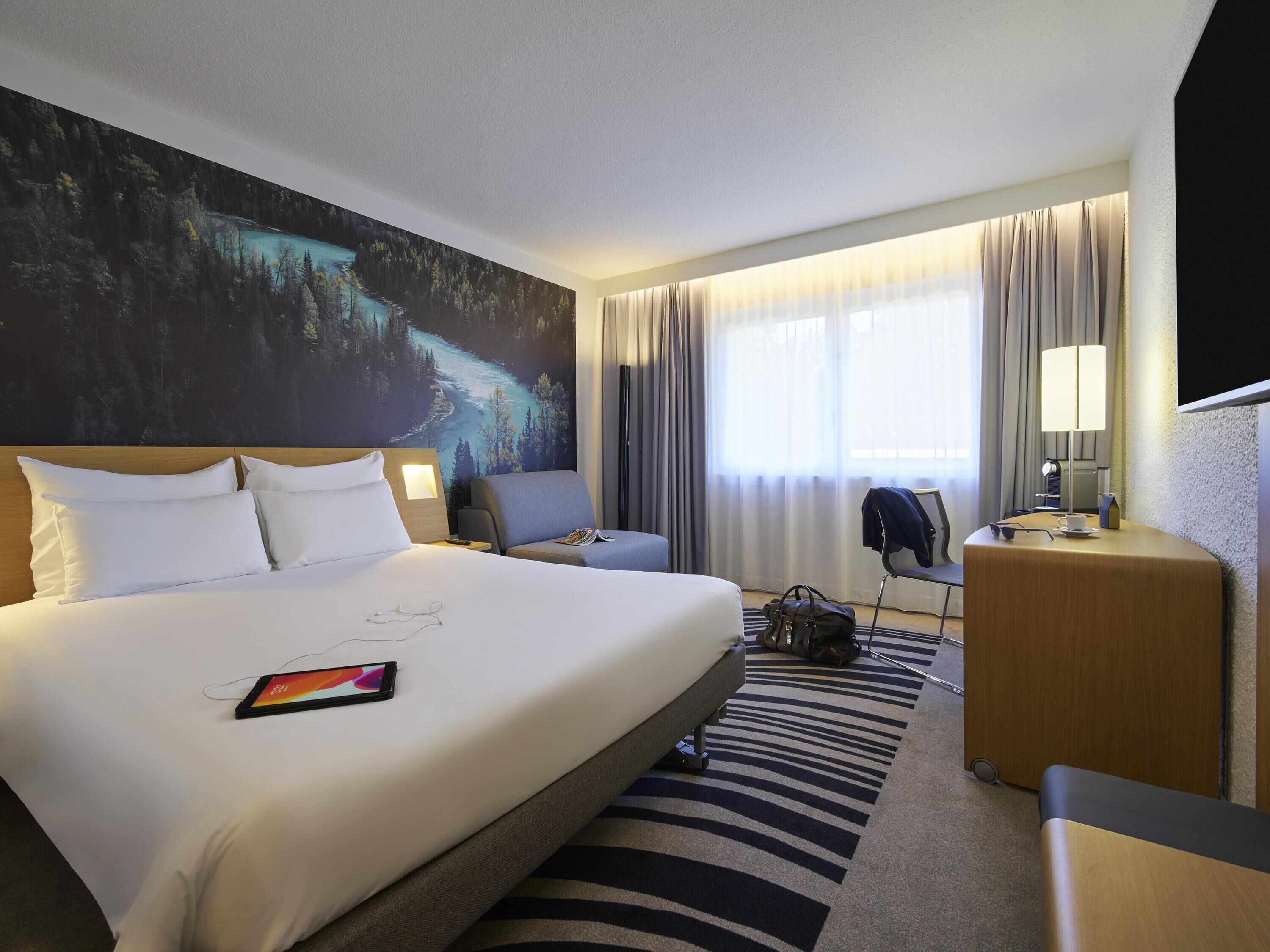 Une chambre d’hôtel avec un grand lit, un bureau, une chaise, une lampe et un tableau représentant une forêt accroché au mur.