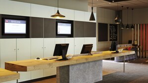 A reception desk with two computer monitors, a wooden counter, and a grey base. There are two pendant lights above the desk. - ibis Strasbourg Centre Historique (Strasbourg)