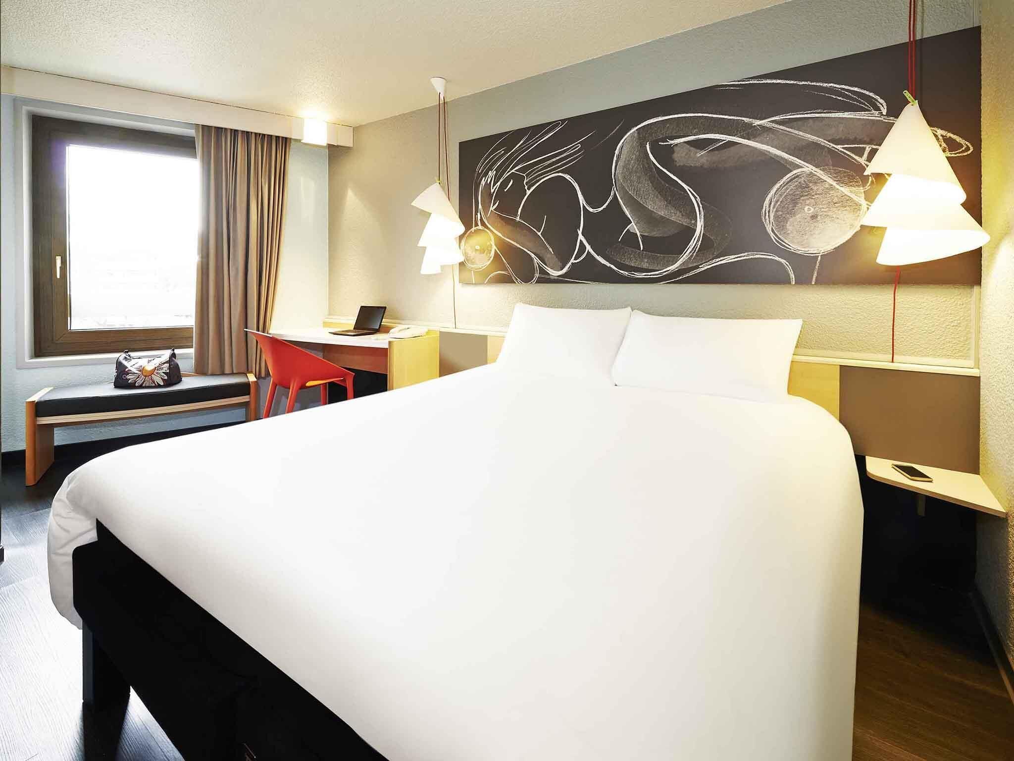 A hotel room with a large bed, a desk, a chair, a window with curtains, and a wall art piece.