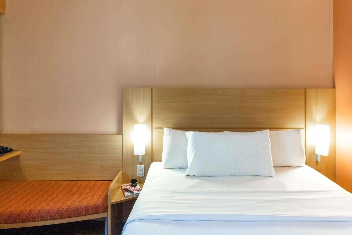 A hotel room with a wooden headboard, two bedside lamps, a white bed, a wooden nightstand, and a red cushioned bench.