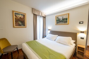 Una habitación de hotel con una cama, una silla, dos cuadros, una mesita de noche y una lámpara.