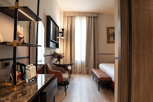 Una camera d'albergo con un letto, una scrivania con una sedia, un tavolino con un vaso di fiori, una televisione a muro e una finestra con tende.