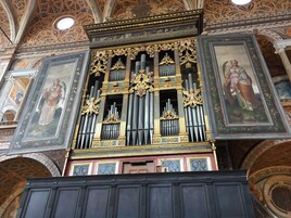 Een sierlijk orgel met gouden details en meerdere pijpen, geplaatst in een rijk versierd interieur met fresco's en architecturale elementen.