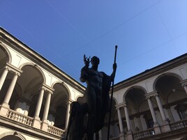 A statue of a muscular figure holding a staff, set against a backdrop of classical architecture with multiple arches and columns.