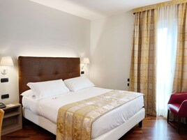 Una camera d'albergo con un letto grande, due lampade ai lati del letto, una poltrona rossa e una finestra con tende.
