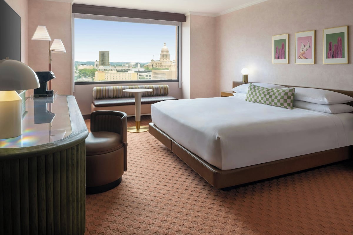 Habitación de hotel con una cama grande, un escritorio con lámpara, una mesita redonda pequeña, un sofá y vistas a un paisaje urbano a través de un ventanal amplio.