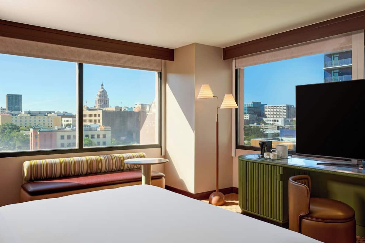 Habitación de hotel con una cama grande, una mesita, un banco, un televisor y vistas a la ciudad.
