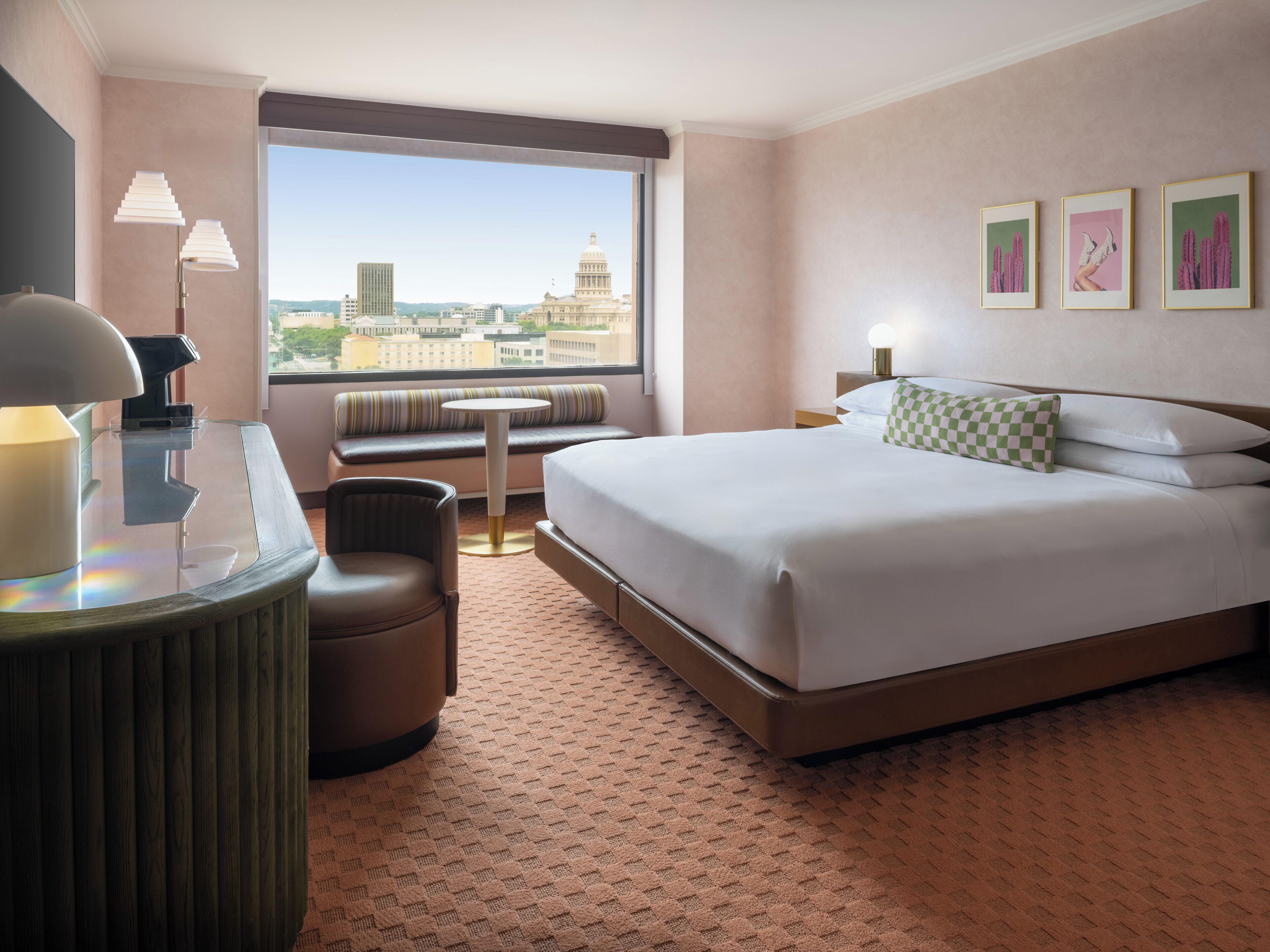 A hotel room with a bed, a desk with a lamp, a chair, a small table, a window with a city view, and three framed pictures on the wall.