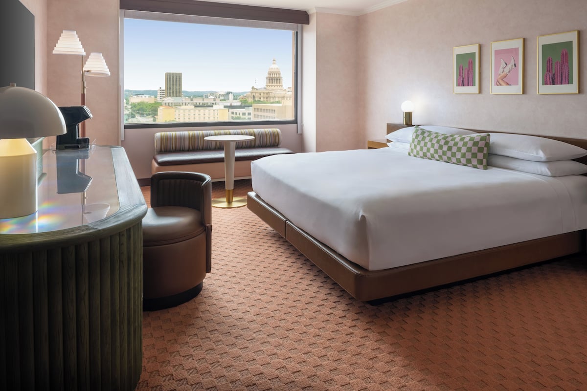 A hotel room with a bed, a desk with a lamp, a chair, a small table, a window with a city view, and three framed pictures on the wall.