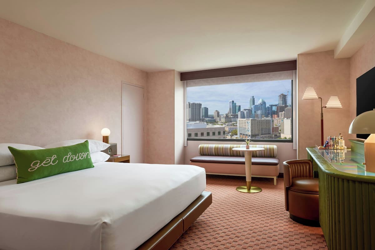 Habitación de hotel con cama, un banco, un escritorio y vistas a un horizonte urbano.
