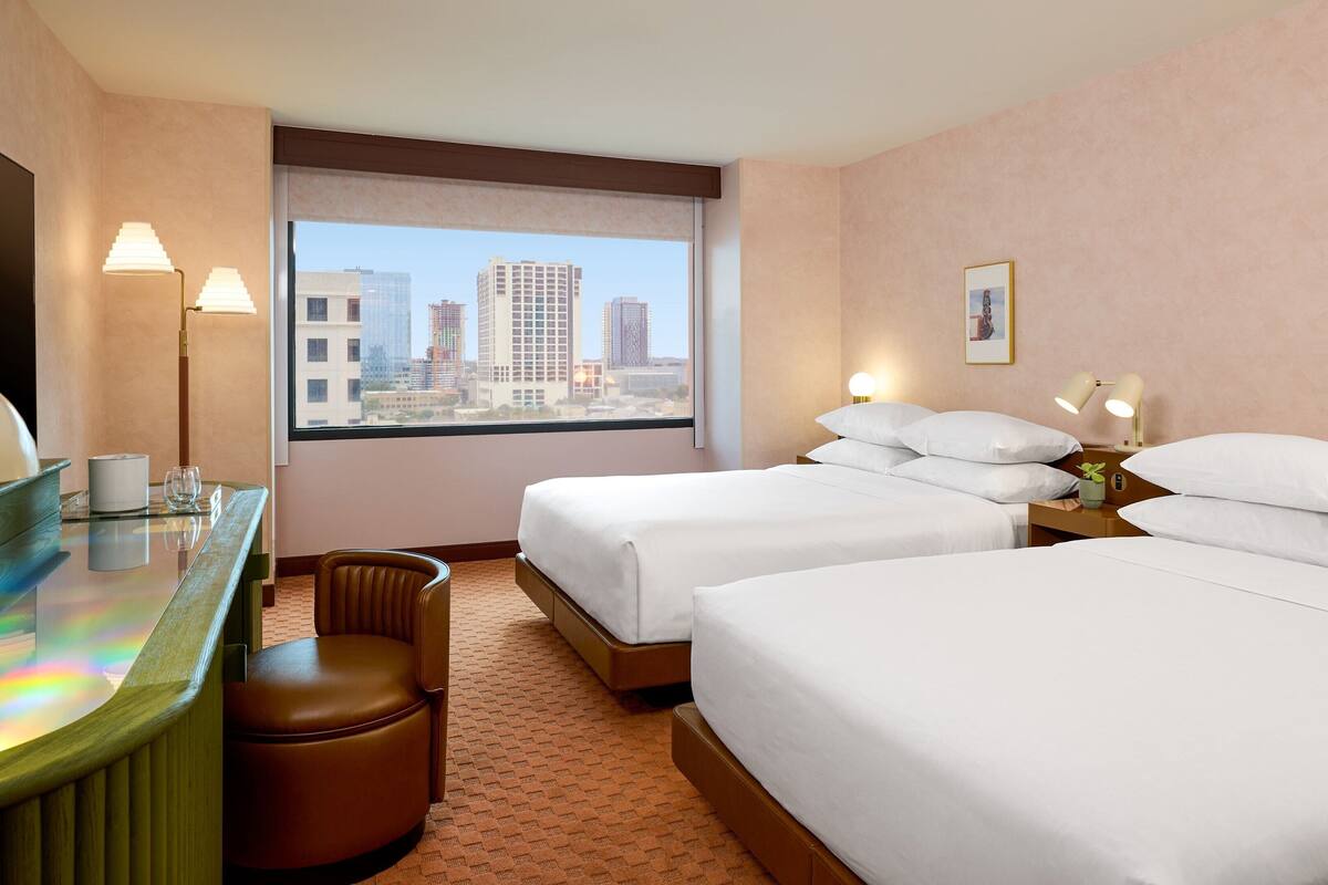 Habitación de hotel con dos camas, un escritorio con silla, vista a la ciudad y un ventanal grande.