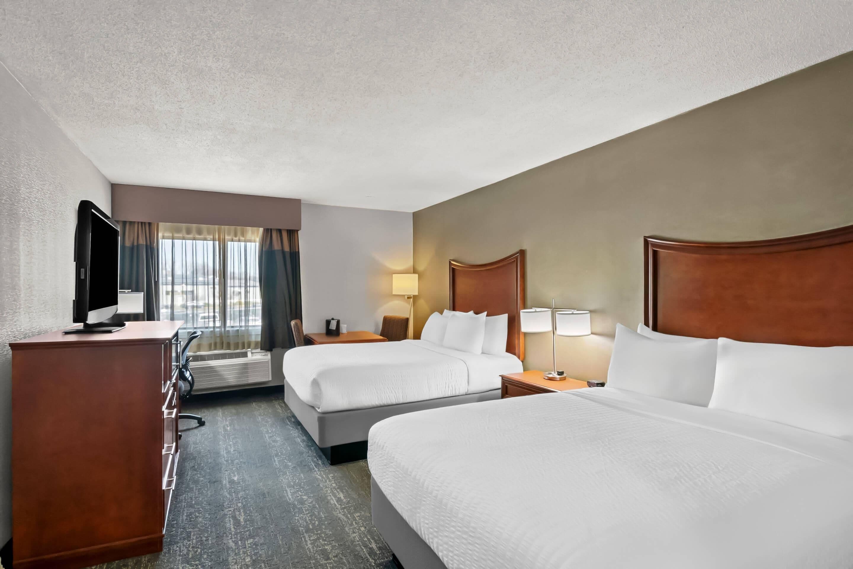 A hotel room with two beds, a desk with a flat-screen TV, a dresser, and a window with curtains.
