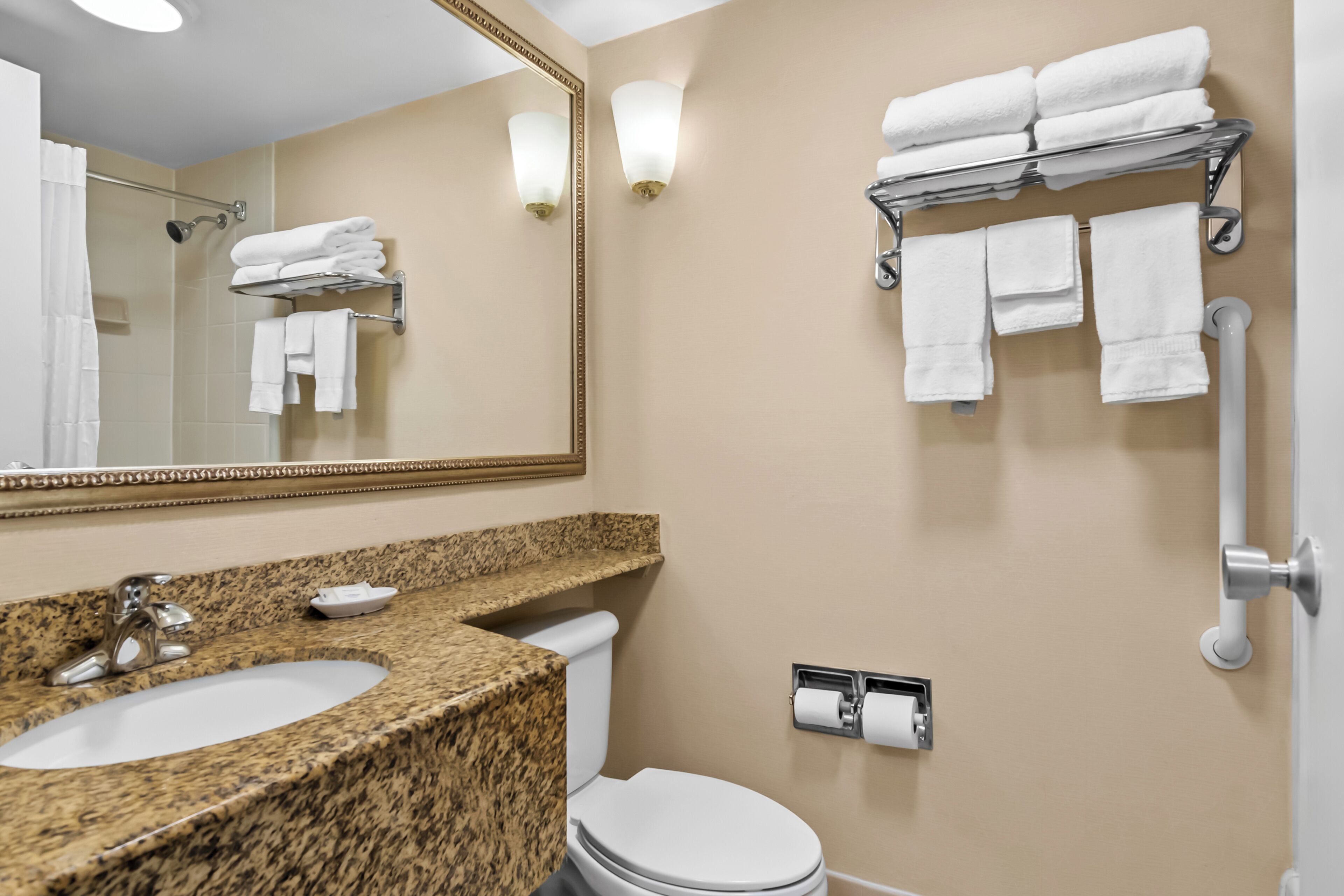 A bathroom with a granite countertop sink, a toilet, a towel rack with folded towels, a mirror with a gold frame, and a wall-mounted toilet paper holder.