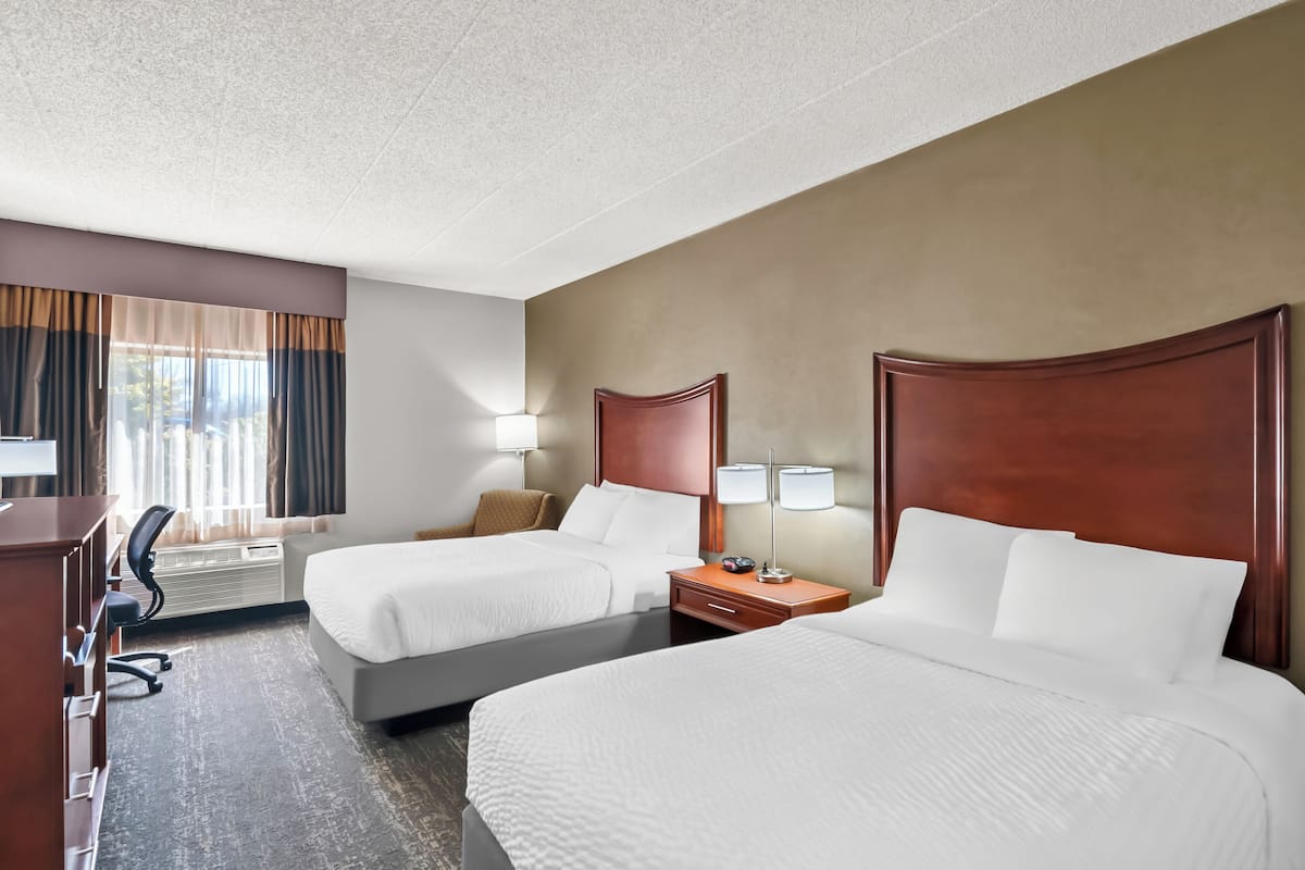 Une chambre d’hôtel avec deux lits, un bureau, une chaise, une lampe et une fenêtre avec des rideaux.