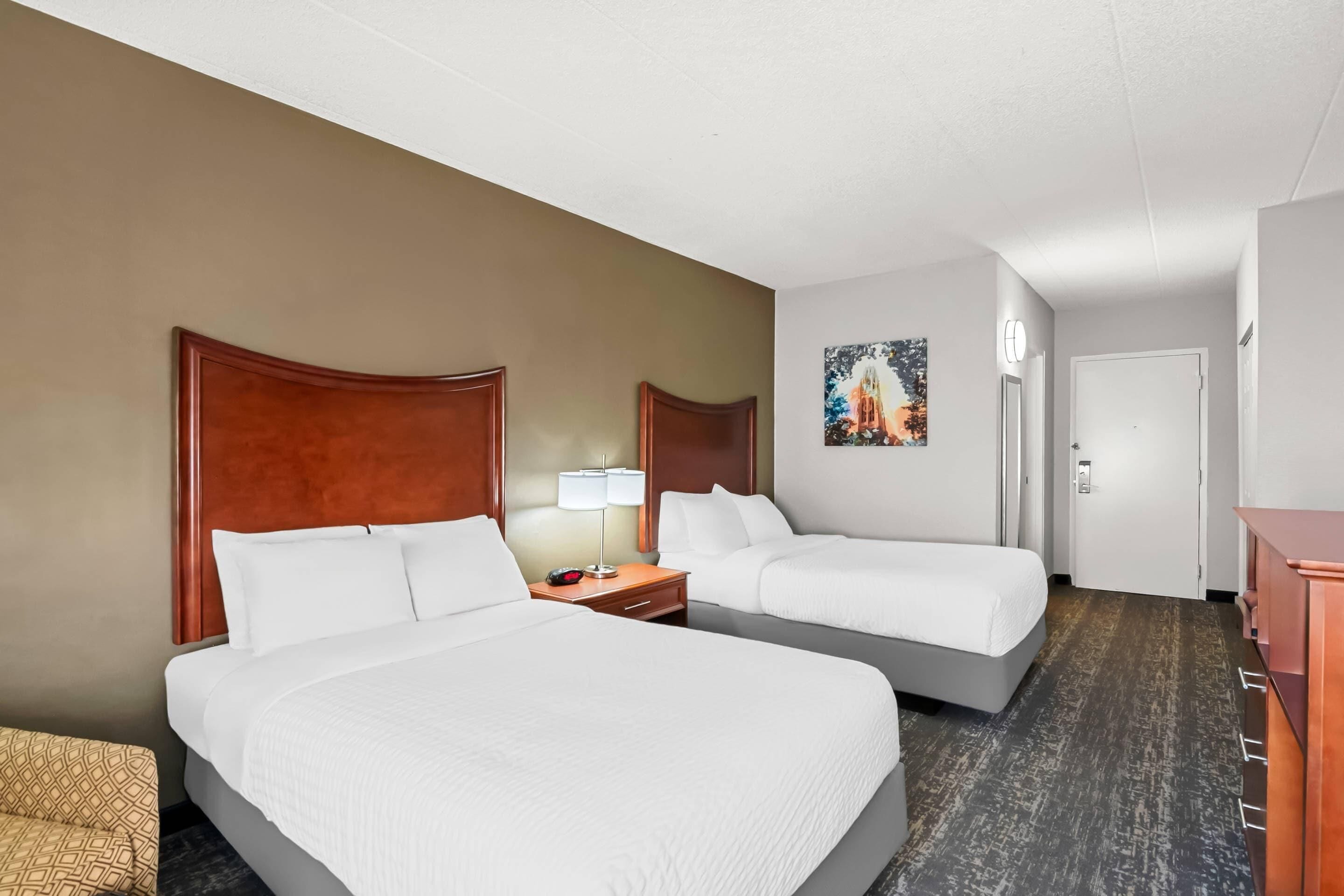 A hotel room with two beds, a nightstand with a lamp, a painting on the wall, and a wooden dresser.