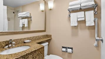 A bathroom with a granite countertop sink, a toilet, a towel rack with folded towels, a mirror, and a wall-mounted toilet paper holder.
