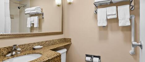 A bathroom with a granite countertop sink, a toilet, a towel rack with folded towels, a mirror, and a wall-mounted toilet paper holder.