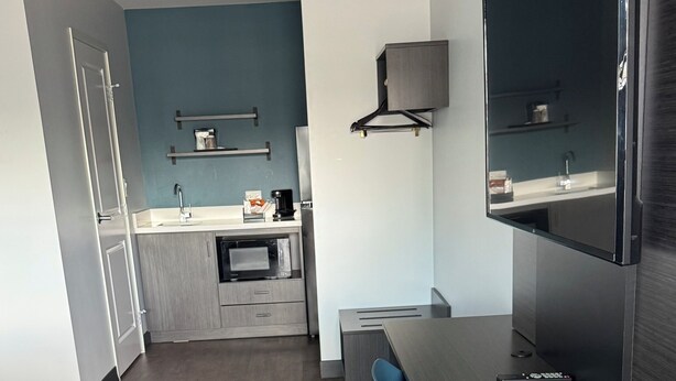 A modern kitchen with a dark countertop, a built-in oven, and a microwave. There is a blue chair and a small table with a remote control on it. - Studio 6 Corona, CA - West (Corona)